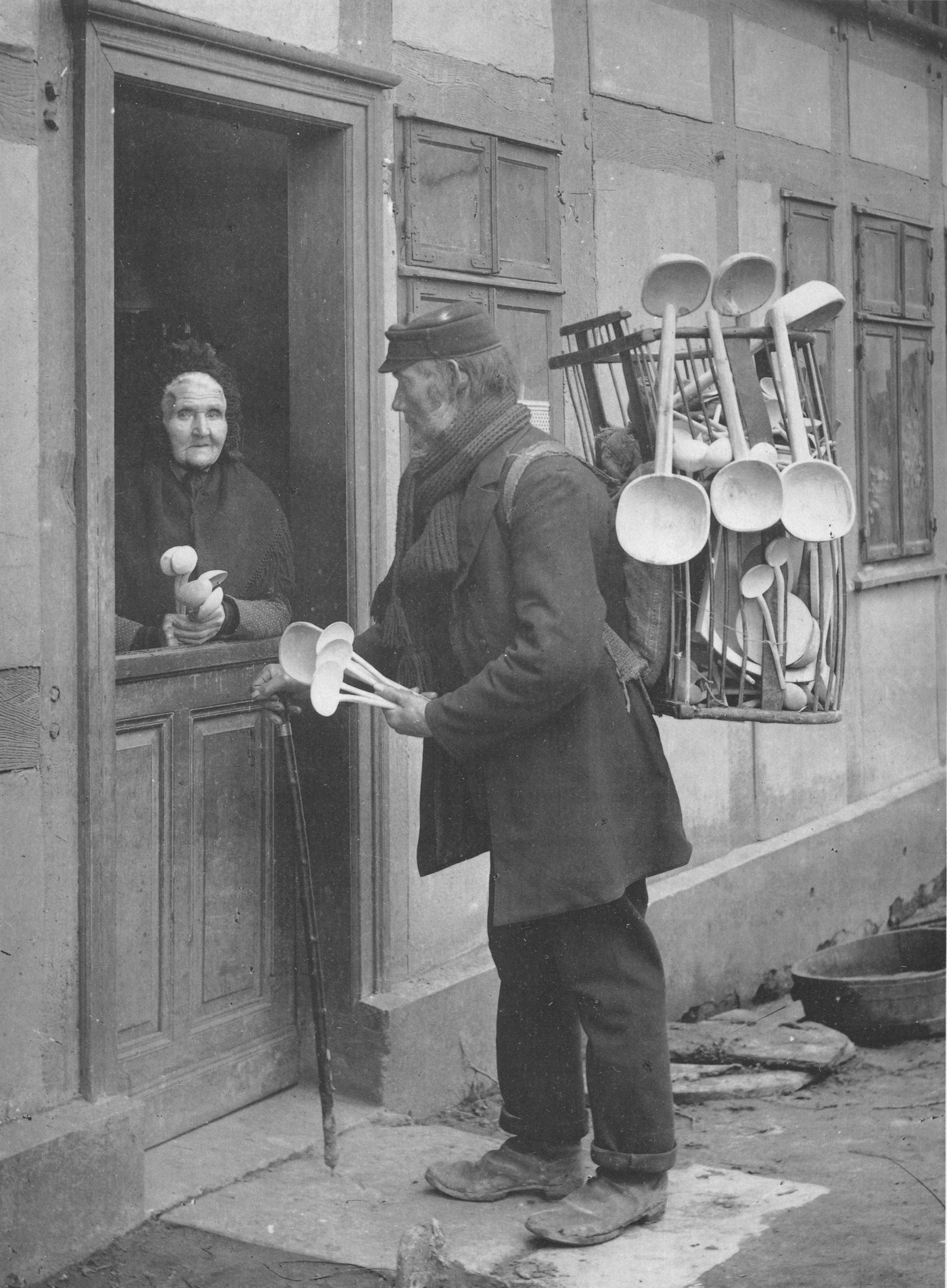 Kiepkerel met houten lepels bij een dame aan de deur, 1905. (Holzwarenhändler, Rahden, 1905). Bron: Dietmar Sauermann, Gerda Schmitz: Alltag auf dem Lande. Auteur: Gottlieb Schäffer (1861-1934). Licentie: Publieke Domein.